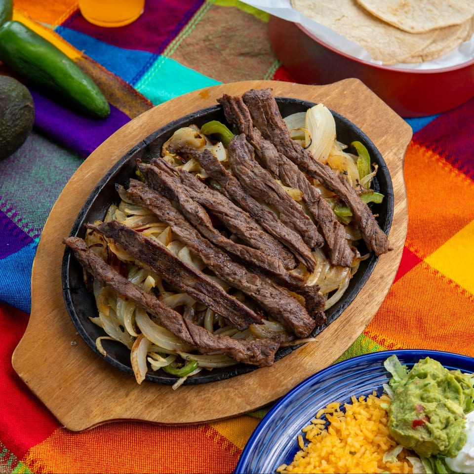 Sizzling steak fajitas with onions and peppers, served with rice, guacamole, and tortillas on a colorful tablecloth.