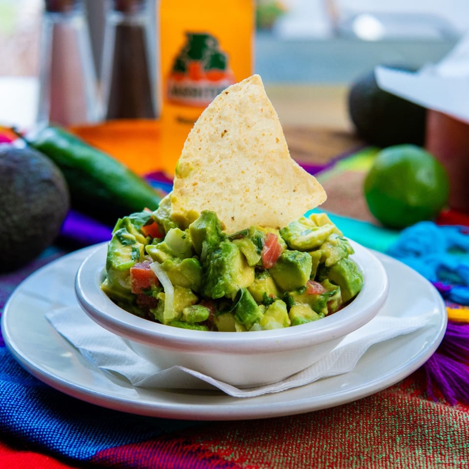 Fresh guacamole with a tortilla chip, surrounded by avocados, lime, and a colorful Mexican blanket.