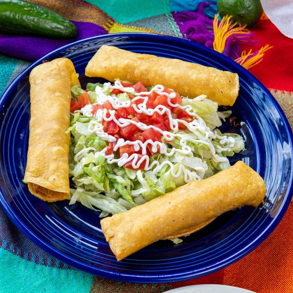 Three golden-brown taquitos with shredded lettuce, diced tomatoes, and sour cream on a blue plate.