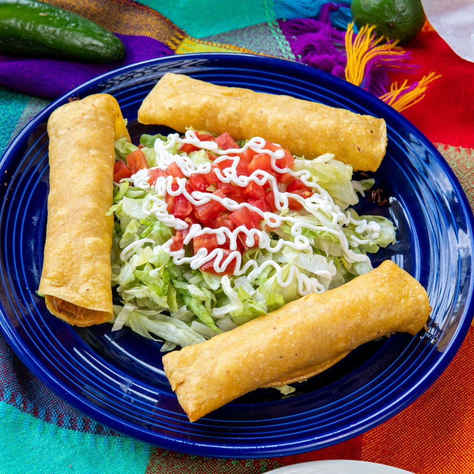 Three golden-brown taquitos with shredded lettuce, diced tomatoes, and sour cream on a blue plate.